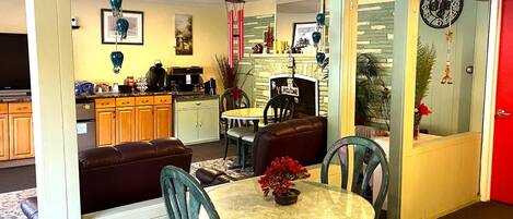 Una sala de estar acogedora con un sofá de cuero marrón, una mesa de centro de vidrio con un centro de mesa de flores rojas y sillas color turquesa. Se ve una cocina con armarios de madera y un refrigerador al fondo.