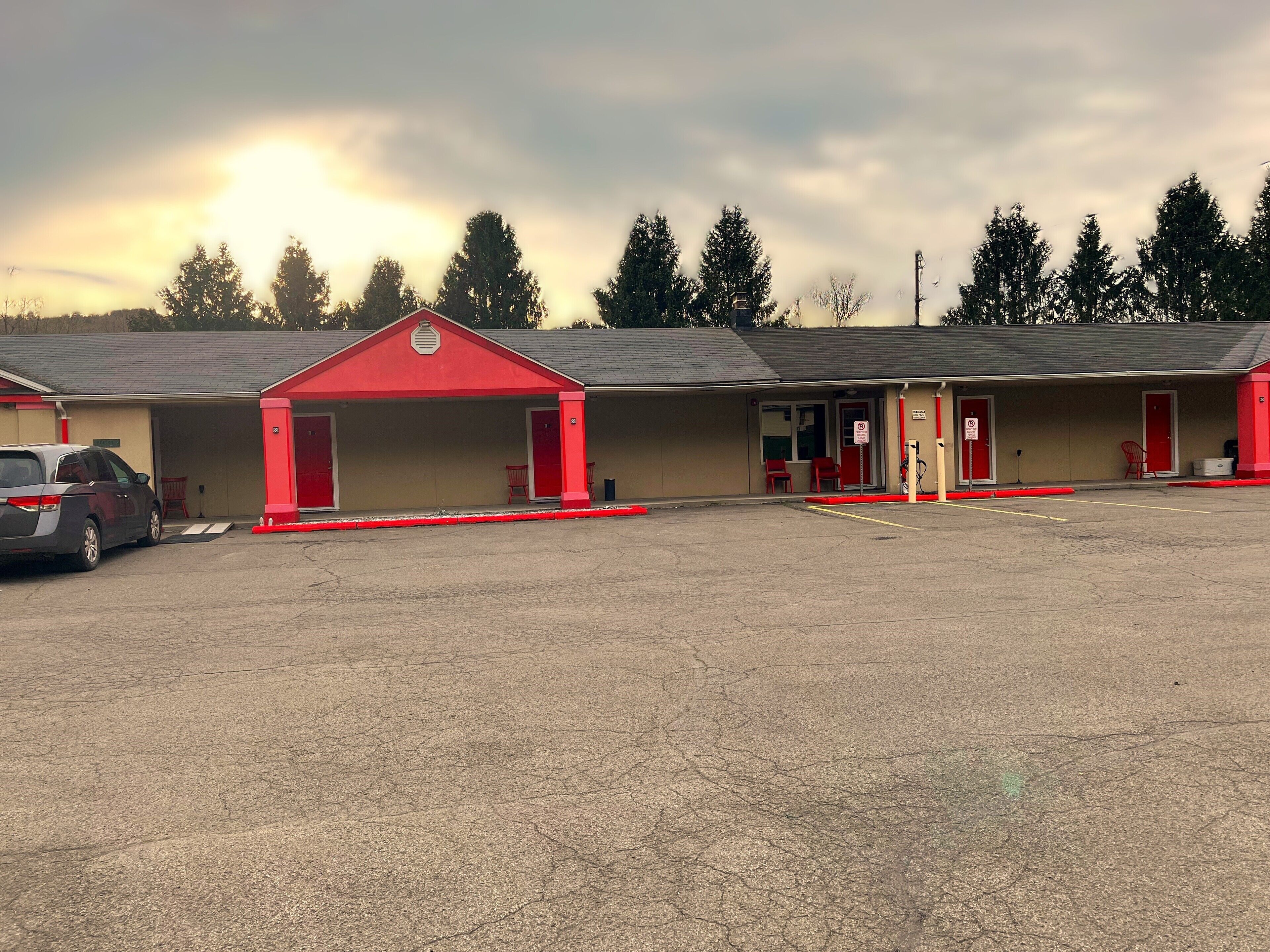 Un motel con entrada roja, un estacionamiento y un hilera de árboles al fondo.