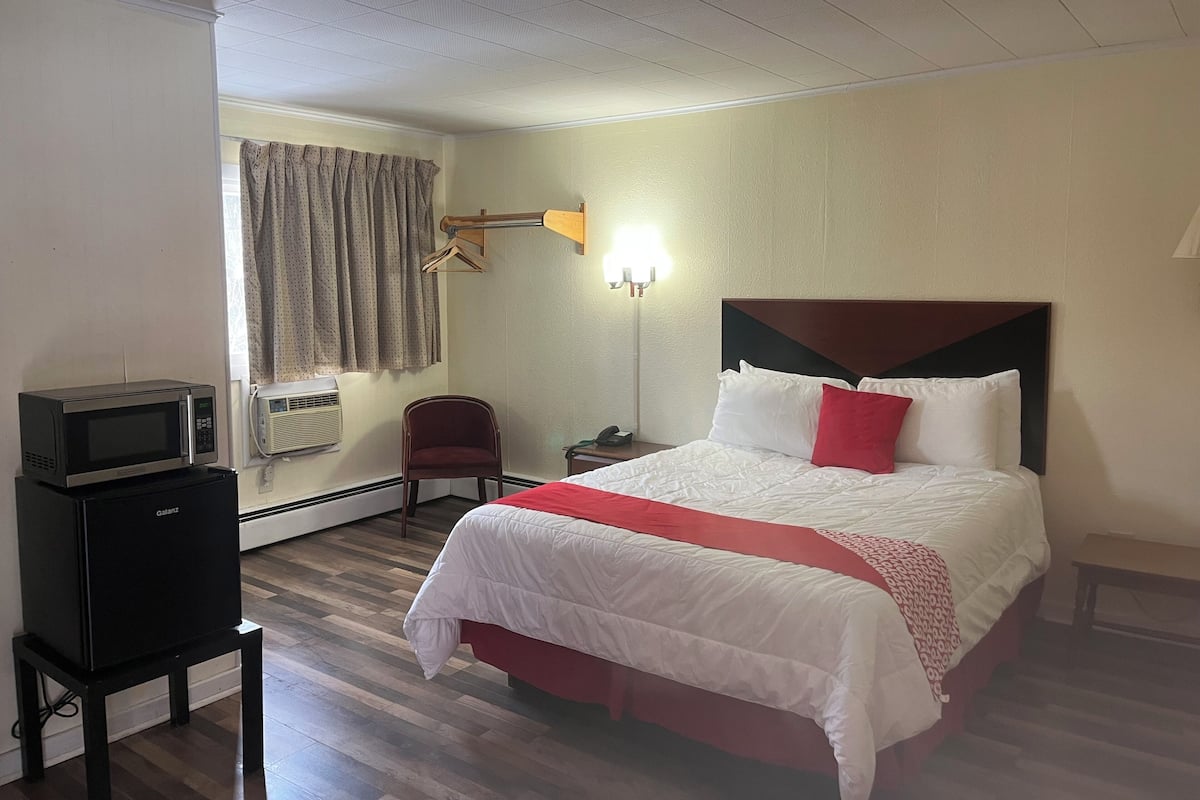 A hotel room with a bed, a desk with a microwave, a chair, a bedside table, and a lamp.