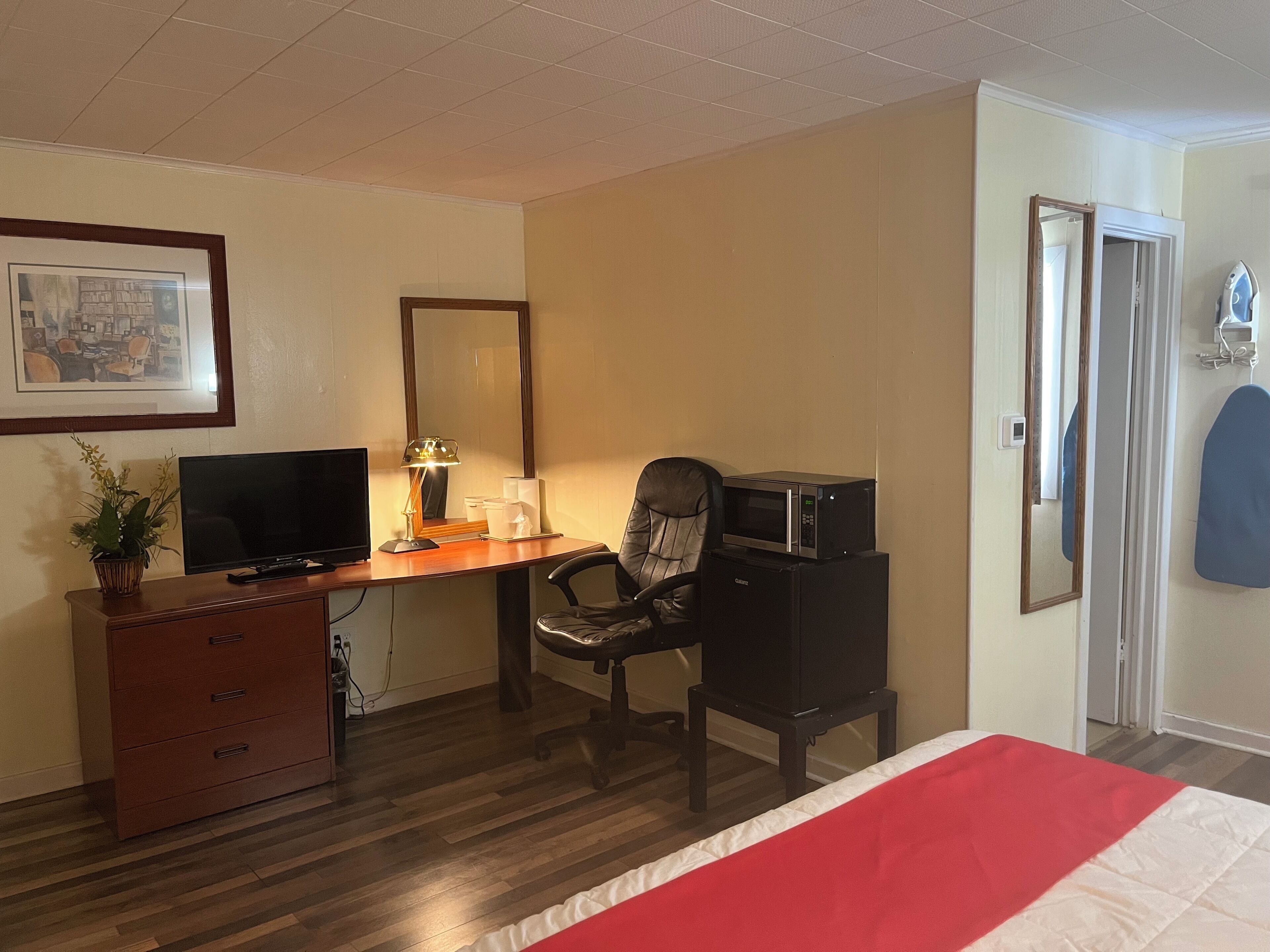 A hotel room with a wooden desk, a chair, a television, a microwave, a dresser, a bed with a red and white bedspread, a framed picture on the wall, and a door with a mirror.