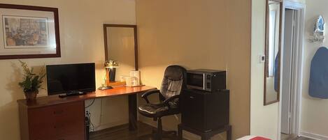 A hotel room with a wooden desk, a chair, a television, a microwave, a dresser, a bed with a red and white bedspread, a framed picture on the wall, and a door with a mirror.