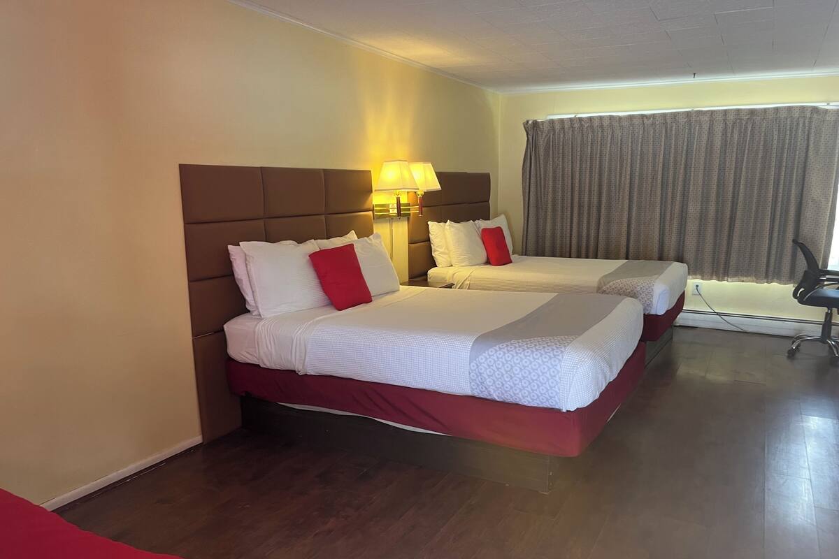 A hotel room with two beds, a wooden floor, a window with curtains, and a desk with a chair.