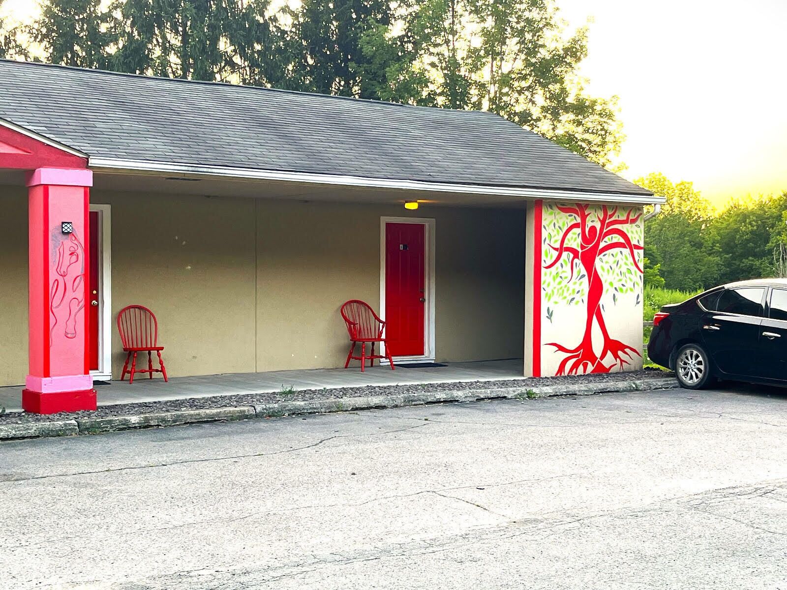 Un edificio con fachada roja y blanca, dos sillas rojas afuera y un mural de un árbol en la pared.
