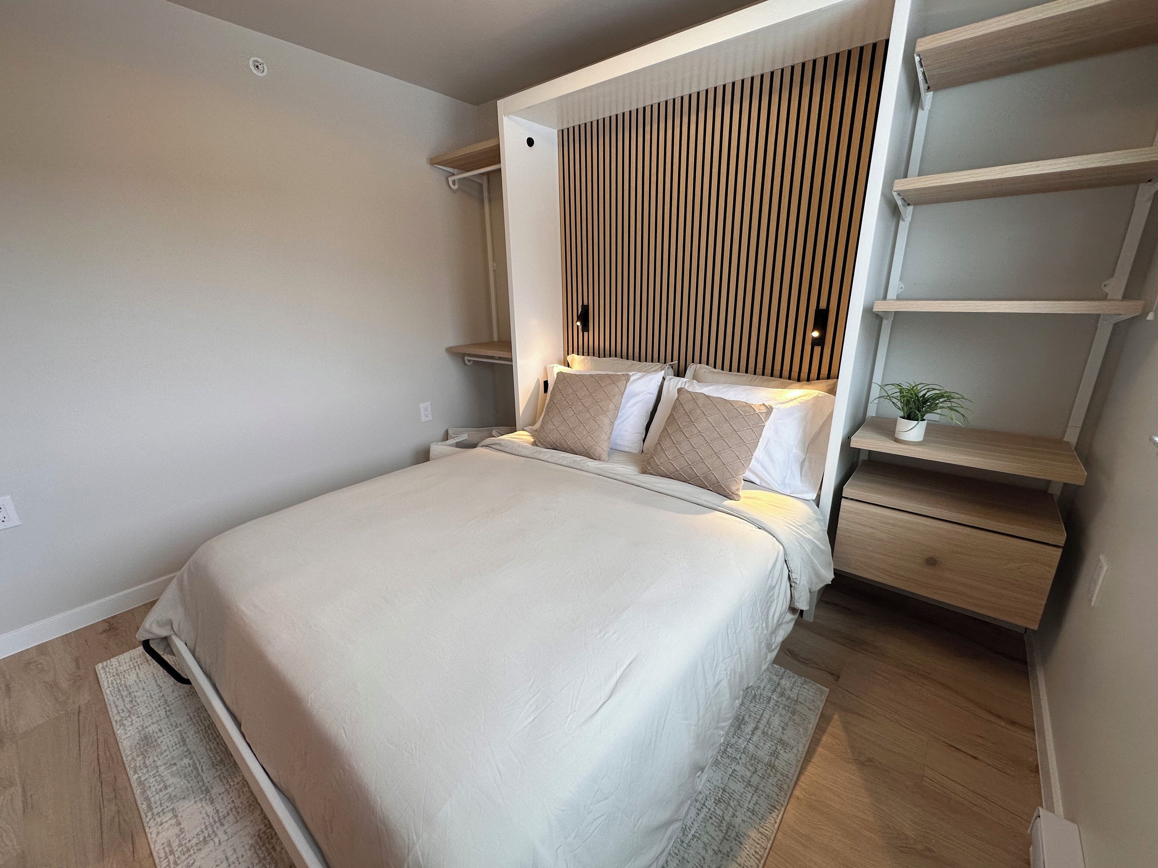 A modern bedroom with a bed, bedside tables, a nightstand, a wall-mounted shelf, and a potted plant.