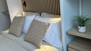 A modern bedroom with a bed, pillows, a nightstand, a plant, and a wooden slatted headboard. - DWELL  in Alaska "BIGGER" Unit o (Fairbanks)