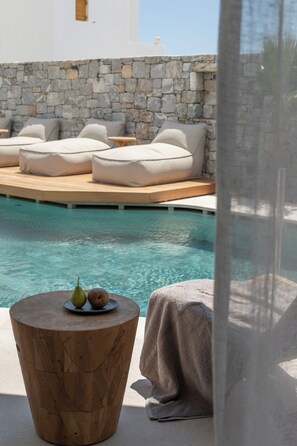 A poolside area with a stone wall, lounge chairs, and a round wooden table with fruits.