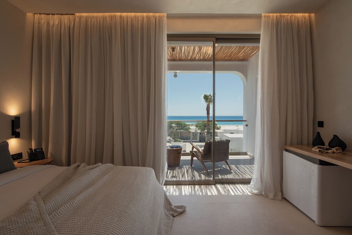 A hotel room with a bed, a desk, a chair, and a balcony with a view of the sea.