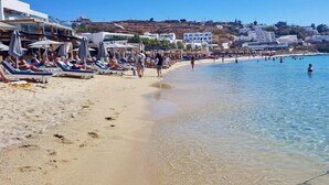 Een strand met ligbedden en parasols, helderblauw water en gebouwen op de achtergrond.