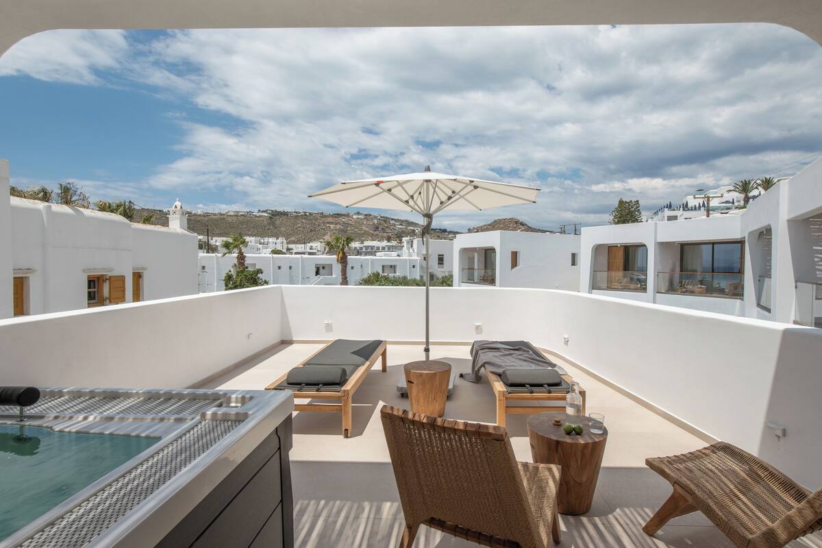A rooftop terrace with lounge chairs, a hot tub, and a view of white buildings and hills.