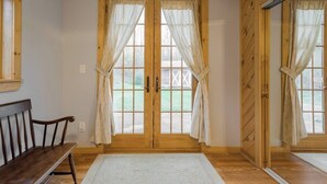 Une pièce avec un plancher en bois, un banc, un tapis blanc et une porte à deux battants avec des rideaux.
