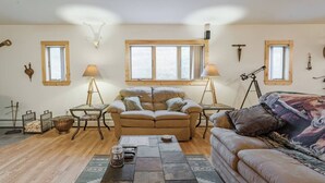 Un salon chaleureux avec un grand canapé, un petit divan, une table basse et deux lampes. On y trouve des fenêtres avec des rideaux, un télescope et un crâne de cerf accroché au mur.