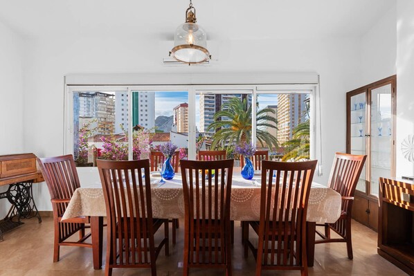 A dining area with a table set for six, wooden chairs, and a view of city buildings through large windows.