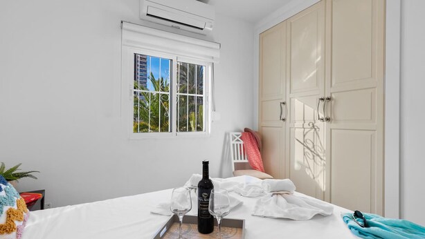 Quarto de hotel com cama, janela vista para palmeiras, aparelho de ar condicionado e guarda-roupa.
