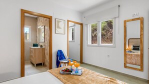 Quarto com cama, penteadeira, cadeira e mesa com bandeja de frutas e copos. Há janelas e um espelho na parede.