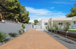 A modern building with a paved courtyard, surrounded by trees and a pool area with lounge chairs.