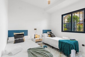 A hotel room with two beds, a window with a view, a bedside table with a lamp, and a small shelf.