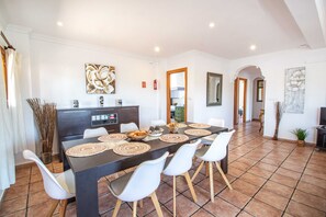 Een eetgedeelte met een lange tafel, witte stoelen en een moderne keuken op de achtergrond.