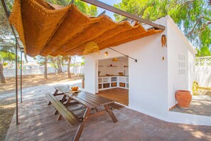 A small, white, single-room structure with a thatched roof, a wooden bench, and a table with fruit.