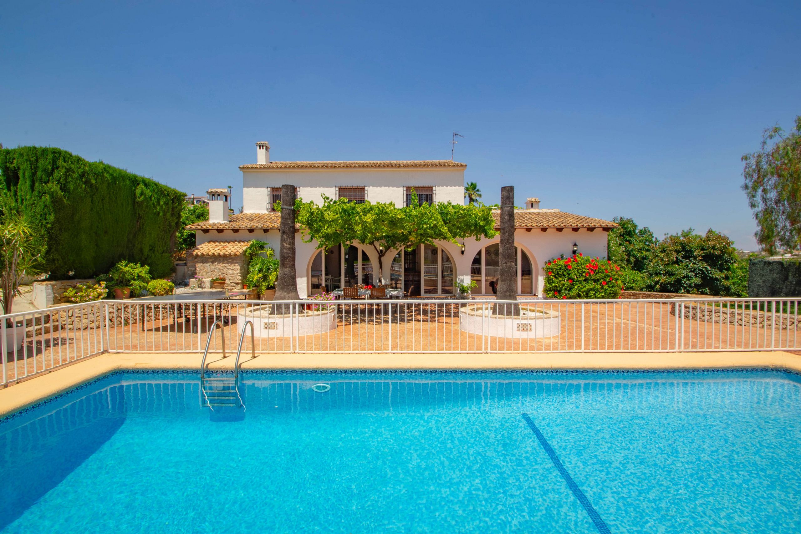 A swimming pool with a white fence, a building with arches, and a clear blue sky.