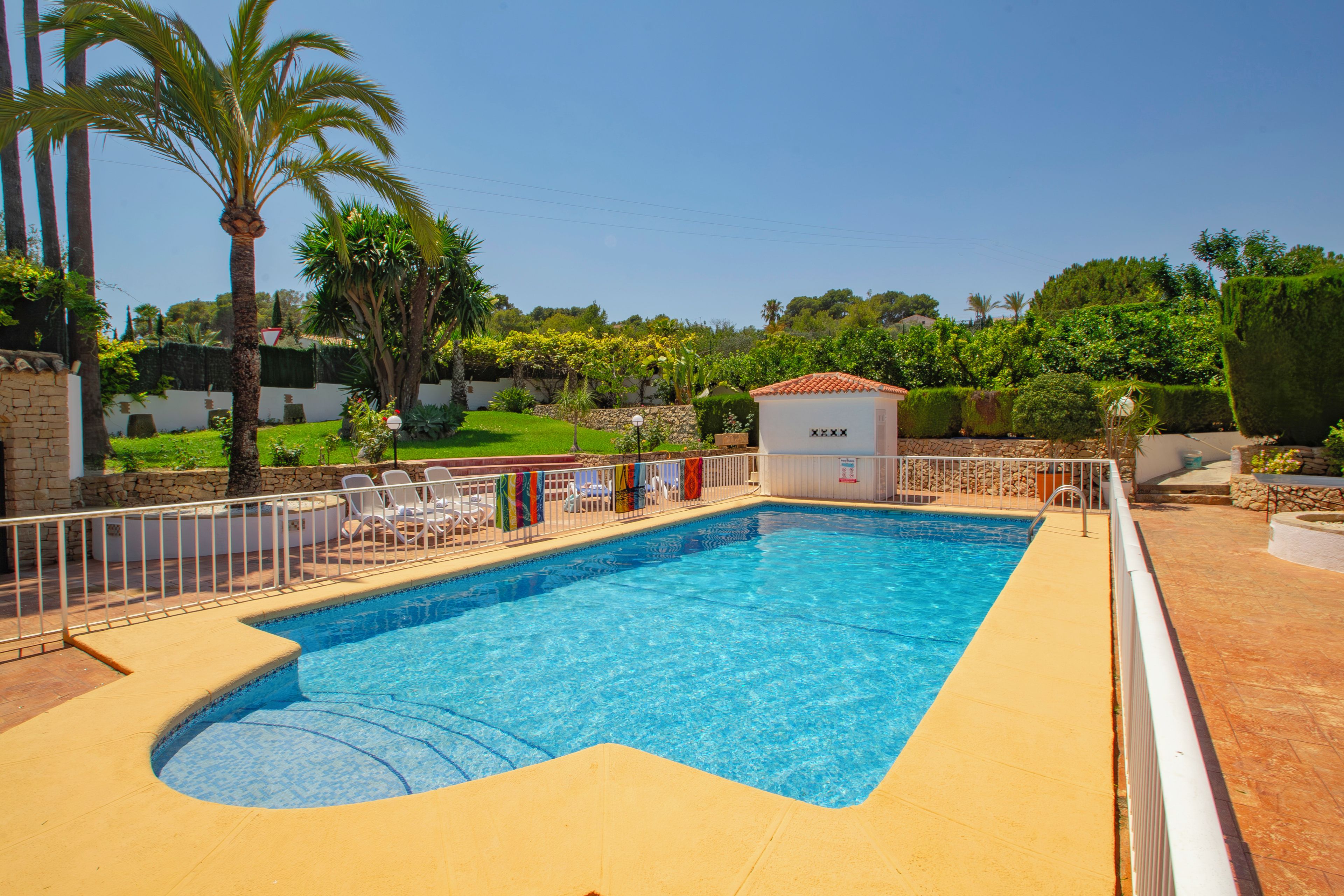 A rectangular swimming pool with a tiled edge, surrounded by a white fence and a few lounge chairs.