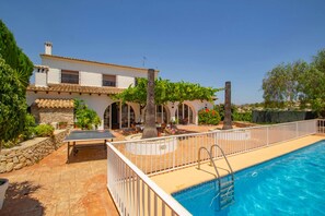 A poolside area with a table tennis table, a pool with a ladder, and a building with arches and a tiled roof.