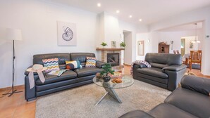 Una sala de estar con sofá, sillón y mesa de centro de vidrio. Se ve una chimenea y un área de comedor al fondo.