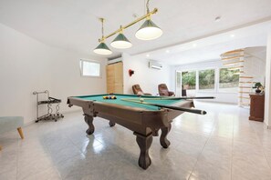 A pool table with a green felt surface, a pool cue, and several balls. There is a bench and a rack of pool cues to the left. The room has white walls, a window, and a spiral staircase in the background.