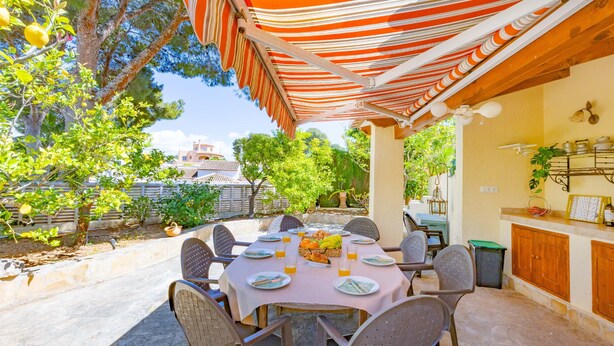 Un área de comedor al aire libre con un toldo a rayas, una mesa preparada para cuatro personas y vistas de la vegetación y edificios circundantes.