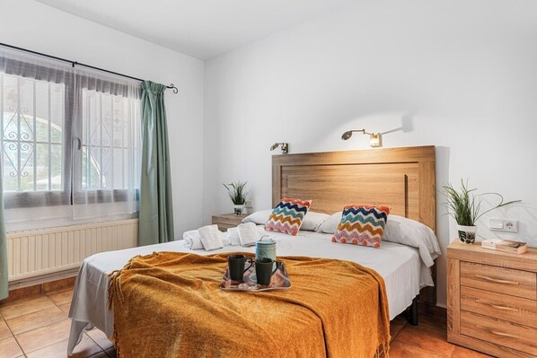 A hotel room with a bed, bedside tables, a nightstand, a window with curtains, and a wall-mounted light fixture.
