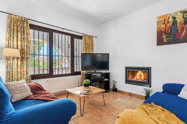 Ein Wohnzimmer mit Fernseher, Kamin, blauen Sofas und einem Fenster mit Vorhängen.