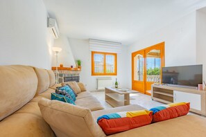A living room with a beige sofa, a wooden coffee table, a television, and a window with orange blinds. - Villa Mona - PlusHolidays (Calpe)