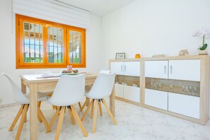 A dining area with a wooden table and white chairs, a white cabinet with glass doors, and a window with wooden frames. - Villa Mona - PlusHolidays (Calpe)