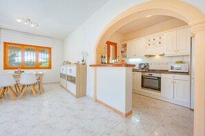 A modern kitchen with white cabinetry, a central island, and a dining area with a table and chairs. - Villa Mona - PlusHolidays (Calpe)