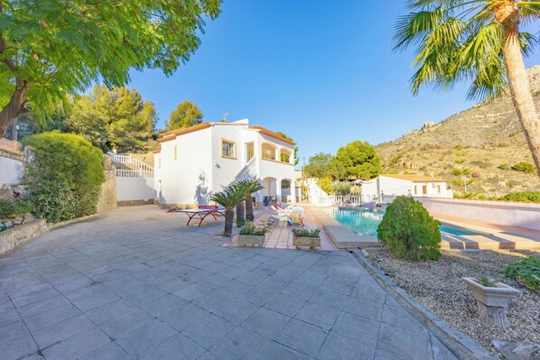 A white villa with a pool, surrounded by trees and a hilly landscape.