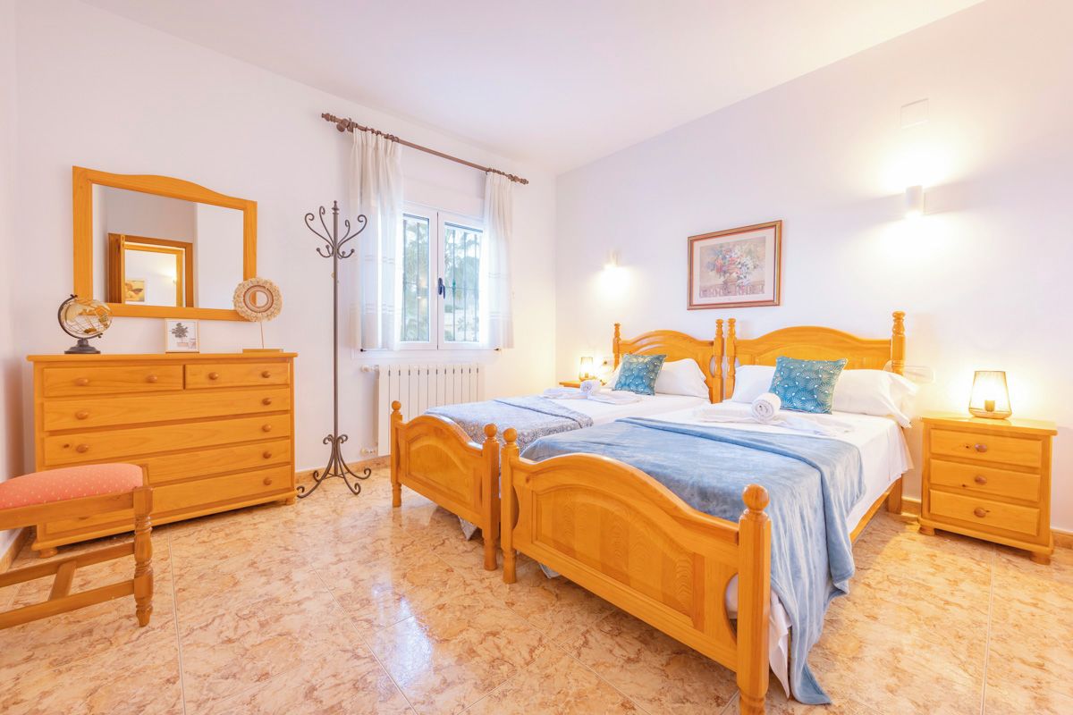 Ein Schlafzimmer mit einem hölzernen Bett, einem Schminktisch, einem Stuhl, einem Nachttisch mit Lampe und einem fenster mit Vorhängen.