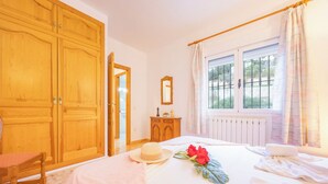 Quarto de hotel com um guarda-roupa de madeira grande, cama com colcha branca, mesa de cabeceira com abajur e janela com cortinas.