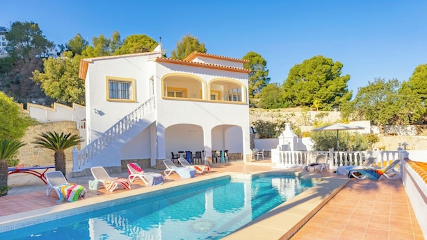 Uma área à beira da piscina com um prédio branco, piscina e espreguiçadeiras.