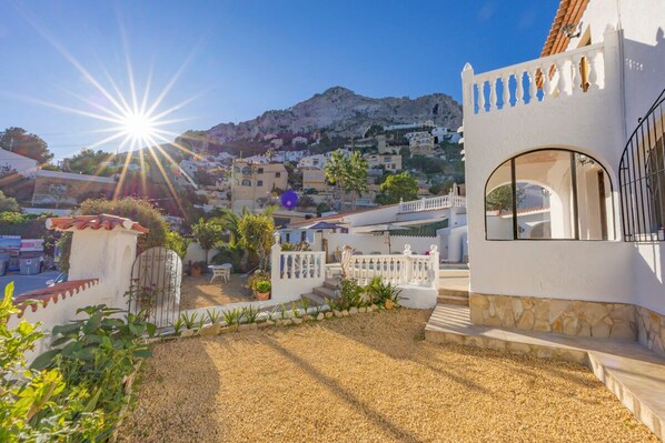 A residential area with a white building featuring a balcony, a gravel driveway, and a mountainous backdrop.