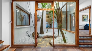 Un interno moderno con una porta in vetro che si apre su un sentiero in pietra, gradini in legno e vista su un albero e un edificio esterni.