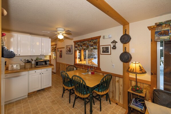 A cozy kitchen with white cabinets, a wooden dining table with chairs, a ceiling fan, and a window with a view of trees.