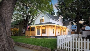 A two-story house with a porch, a flag, and a white picket fence. - Dwntwn CDA Historic Remodel - Walk to Lake with Hot Tub! (Coeur d'Alene)
