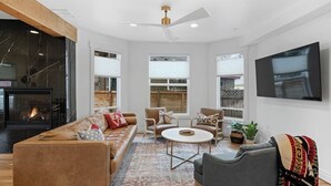A modern living room with a fireplace, a ceiling fan, and a large TV mounted on the wall. - Dwntwn CDA Historic Remodel - Walk to Lake with Hot Tub! (Coeur d'Alene)