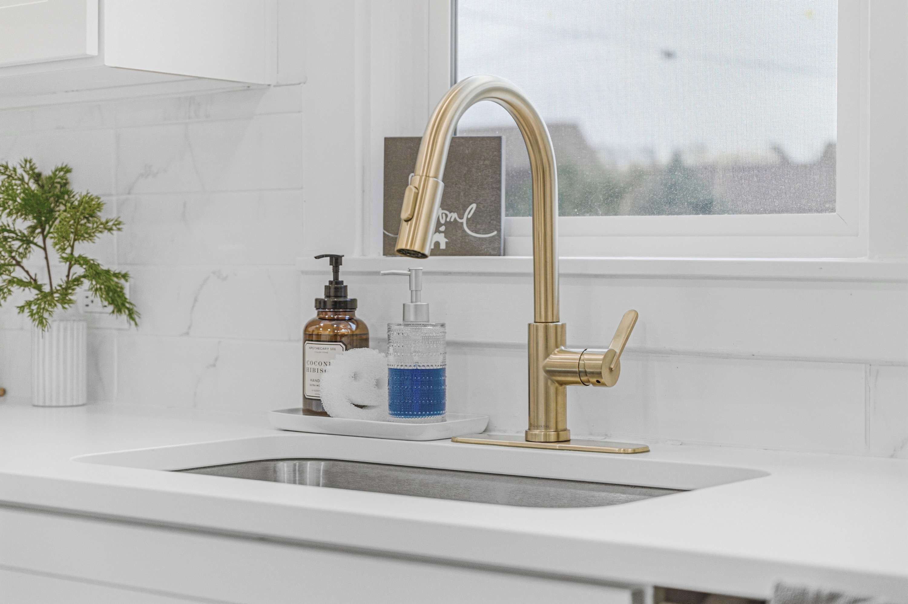 A kitchen with a white countertop, a gold faucet, a soap dispenser, and a glass of blue liquid.