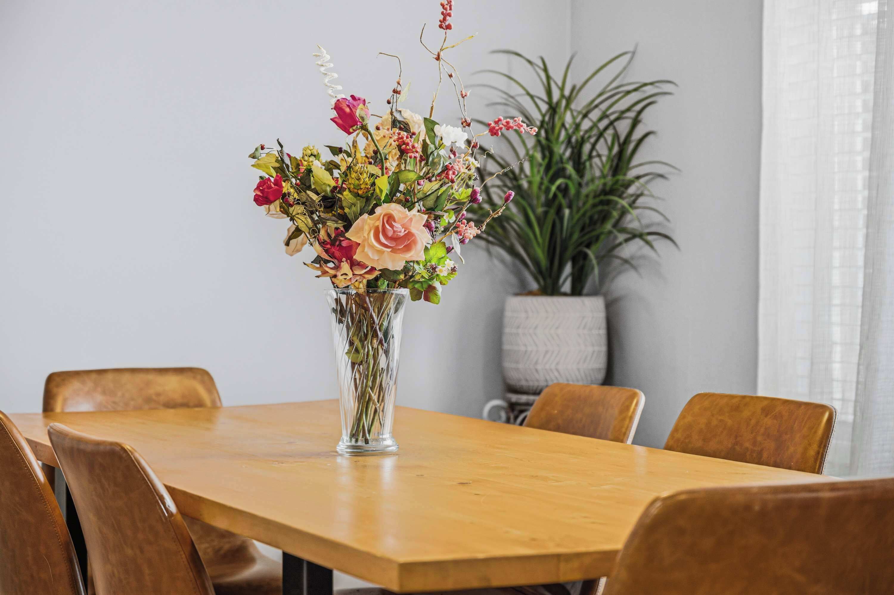Een eettafel met stoelen, een bloevas en een plant in een pot op de achtergrond.
