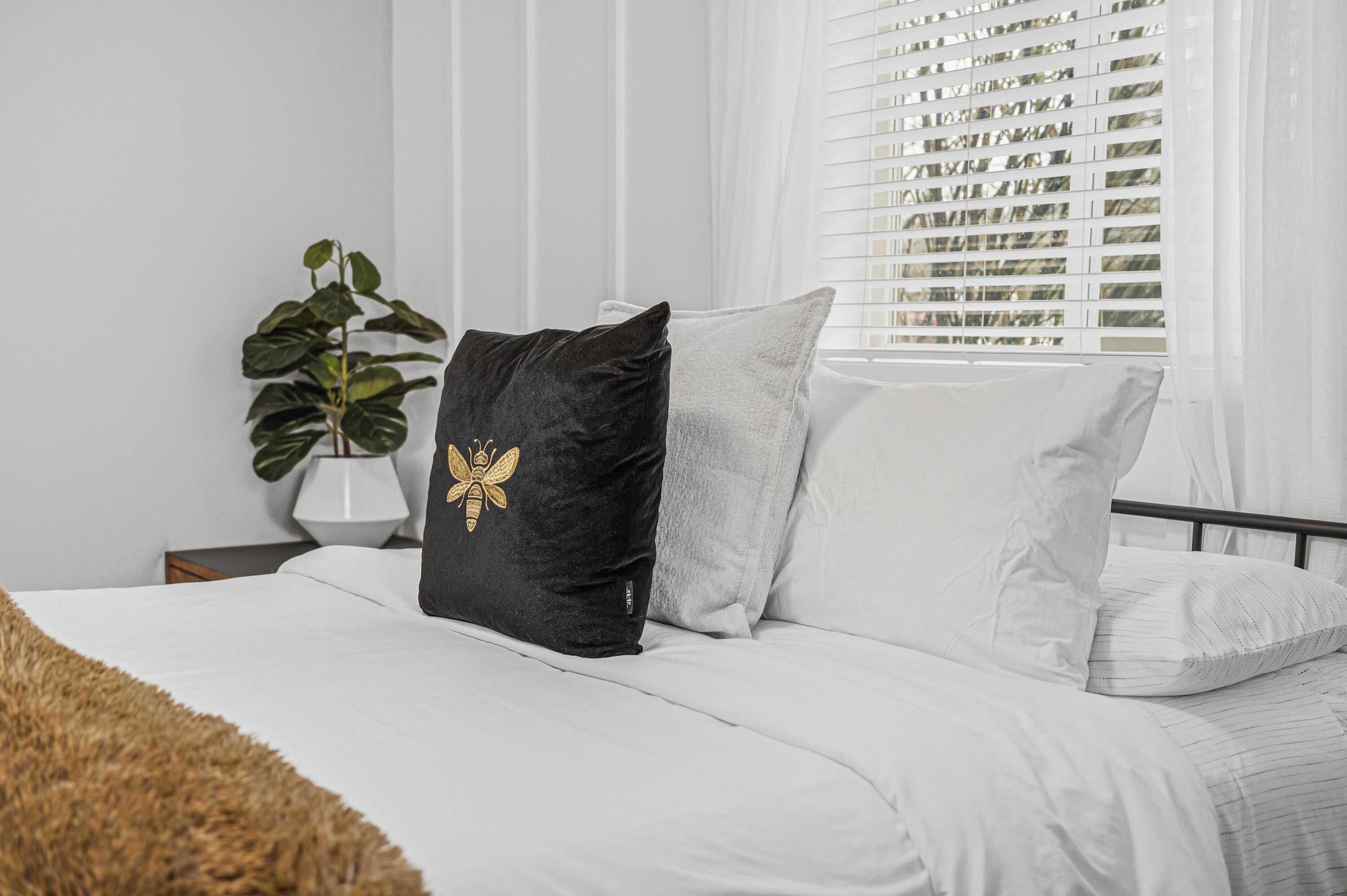 A bed with white bedding, a black pillow with a gold design, and a grey pillow. There is a plant in a white pot on a bedside table.
