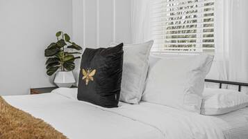 A bed with white bedding, a black pillow with a gold design, and a grey pillow. There is a plant in a white pot on a bedside table.