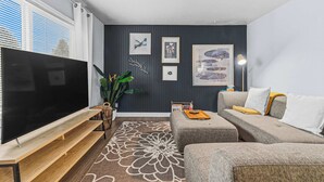 Un salon doté d’un téléviseur à écran plat, d’un canapé gris, d’un tapis à motifs et d’œuvres d’art accrochées au mur.
