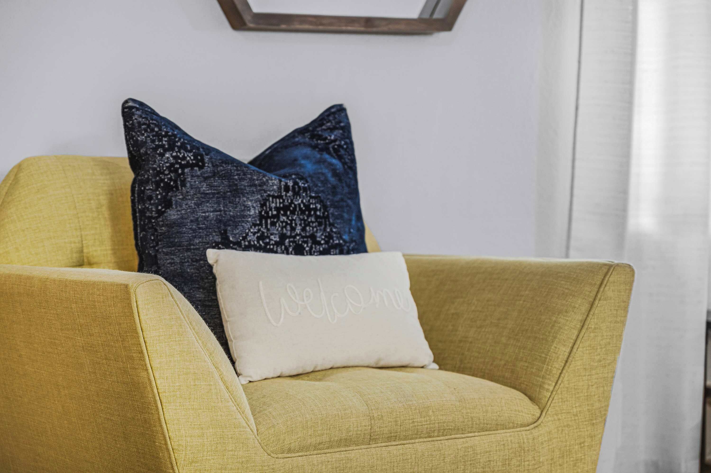 Een gele fauteuil met een kussen waarop 'Welkom' staat en een donkerblauwe kussen met een reliëfpatroon.