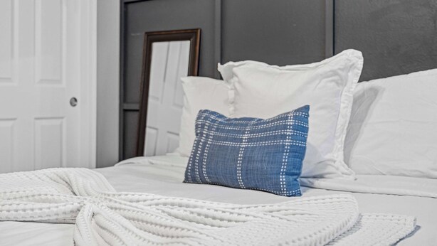 Un lit avec une literie blanche, un oreiller bleu et un plaid blanc.