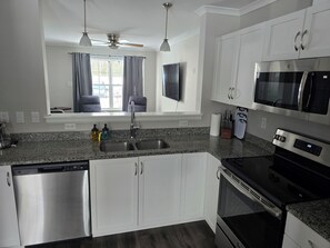 A modern kitchen with white cabinets, stainless steel appliances, a granite countertop, and a window with curtains. - Acadia Villas!  1A Olivia.  A community nestled in the woods. (Trenton)